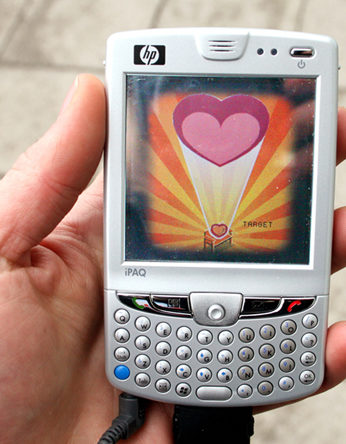 a PDA phone with a heart graphic on the screen held in someone's hand with the street behind