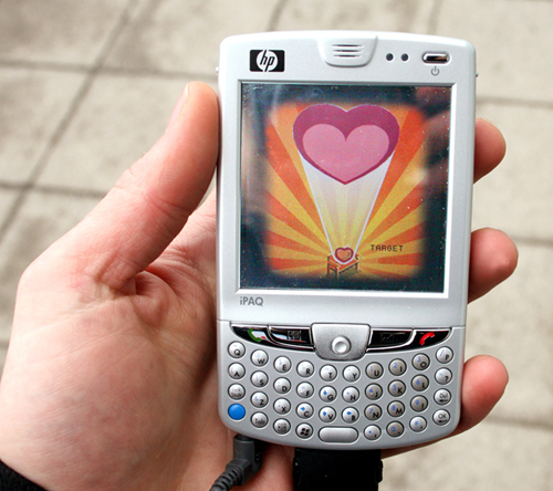 a PDA phone with a heart graphic on the screen held in someone's hand with the street behind