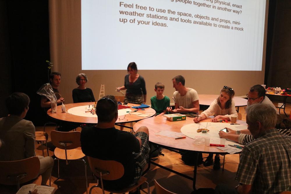 Rachel in front of a large screen talking to people sitting around a table with paper and materials on the table