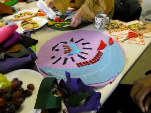 A circular artwork madefrom brightly coloured felt, with plates for biscuits, scissors and glue on the table.