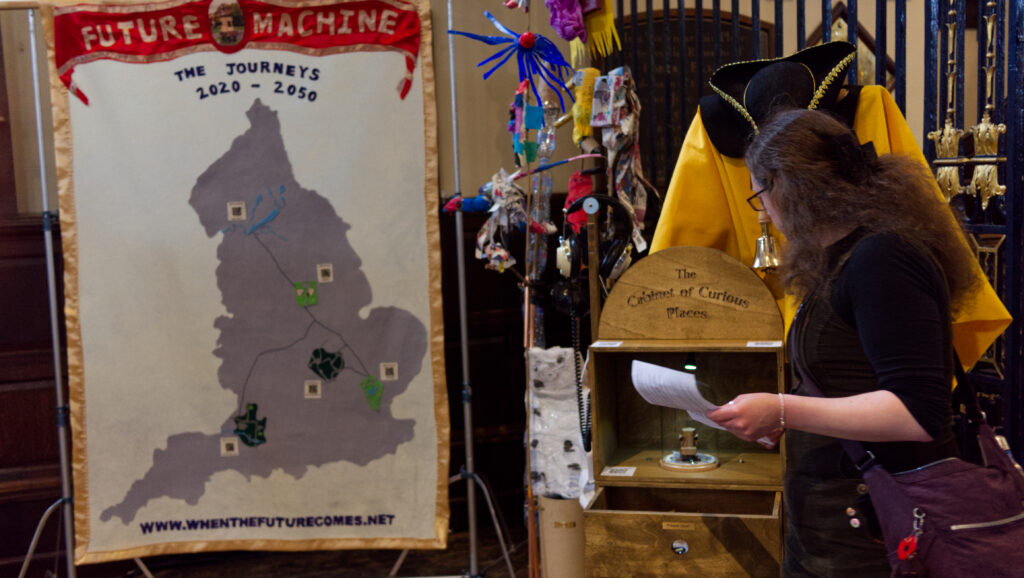 A banner with Future Machine - The Journeys 2020 - 2050 and a map of England with QR codes and symbols for each place where Future Machine visits. Alongside someone looking at the Cabinet of Curious Places