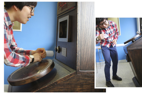 Two images - the left images is of a man turning a wooden circular handle on an upright wooden machine, looking at a screen with a dial below it and a light up sign above it. In the right image the man is bent down reading from a small piece of paper