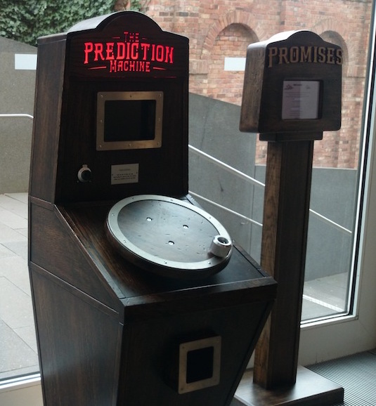Two artworks made of wood. The Prediction Machine has the name lit up in red at the top, a rectangular screen, a dial, a circular hand crank and a hole below, made of dark wood with steel surrounds for each element. The Promises Machine is a wooden stand with a screen at the top.