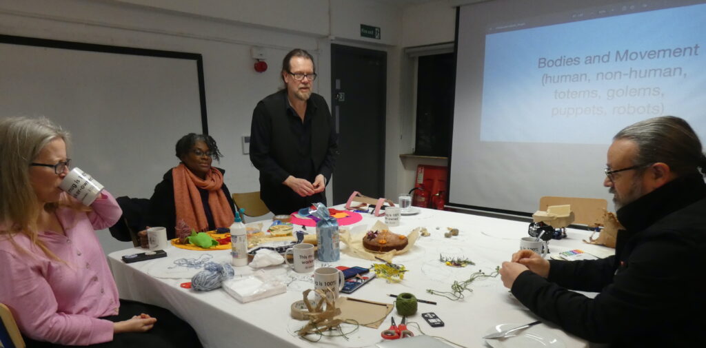 4 people sitting around the table with a projected slide behind them showing the words Bodies and Movement and a table with cake, cups, coloured felt, scissors, cake and flowers on it