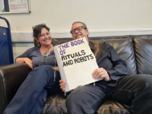 Rachel and Alan sitting on a sofa with The Book of Rituals and Robots