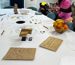 The Convivial Tablecloth with the robots, signs with instructions, a basket of fruit and Rachel's hands showing someone in a pink jacket the table
